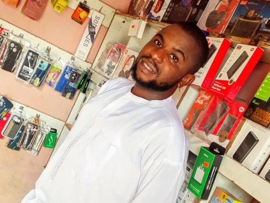 A man in white standing in Achu Owalu&rsquo;s phone-accessories shop, its shelves stocked with phone cases, power banks and earphones