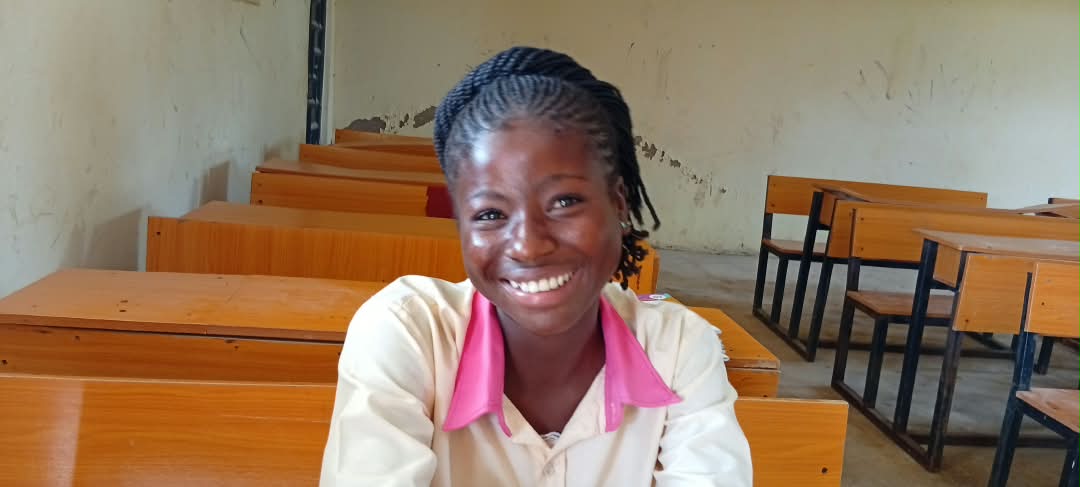A smiling schoolgirl in a cream uniform with a pink collar, at a wooden desk in a classroom