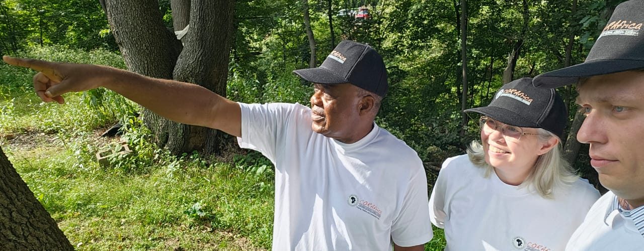 Fr. Peter Abue, in a CORAfrica cap and T-shirt, points into the distance among trees, with Jeannine Goelz and Ethan Suquet beside him
